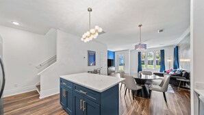 Eine moderne Küche mit einer zentralen Kochinsel, ein Essbereich mit rundem Tisch und Stühlen, sowie eine Wohnbereits mit Sofa und Fernseher.