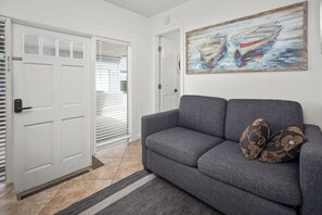 A living room with a grey sofa, two patterned cushions, a painting of boats, and a door with a window. - Mission Beach 1BR · Steps to Bay & Ocean · Water Views · Free Parking (San Diego)