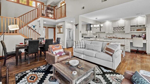Un amplio espacio habitable con una mesa de comedor, sofá y mesa de centro. Una escalera conduce a un nivel superior. La cocina cuenta con gabinetes blancos y una isla central.