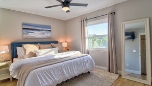 A bedroom with a large bed, a ceiling fan, a window with curtains, a bedside table with a lamp, and a wall-mounted TV. - Stay & Play Tampa- Pool + Mini Golf (Pinellas Park)