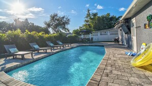 A swimming pool with lounge chairs and a slide, surrounded by a brick patio and trees. - Stay & Play Tampa- Pool + Mini Golf (Pinellas Park)
