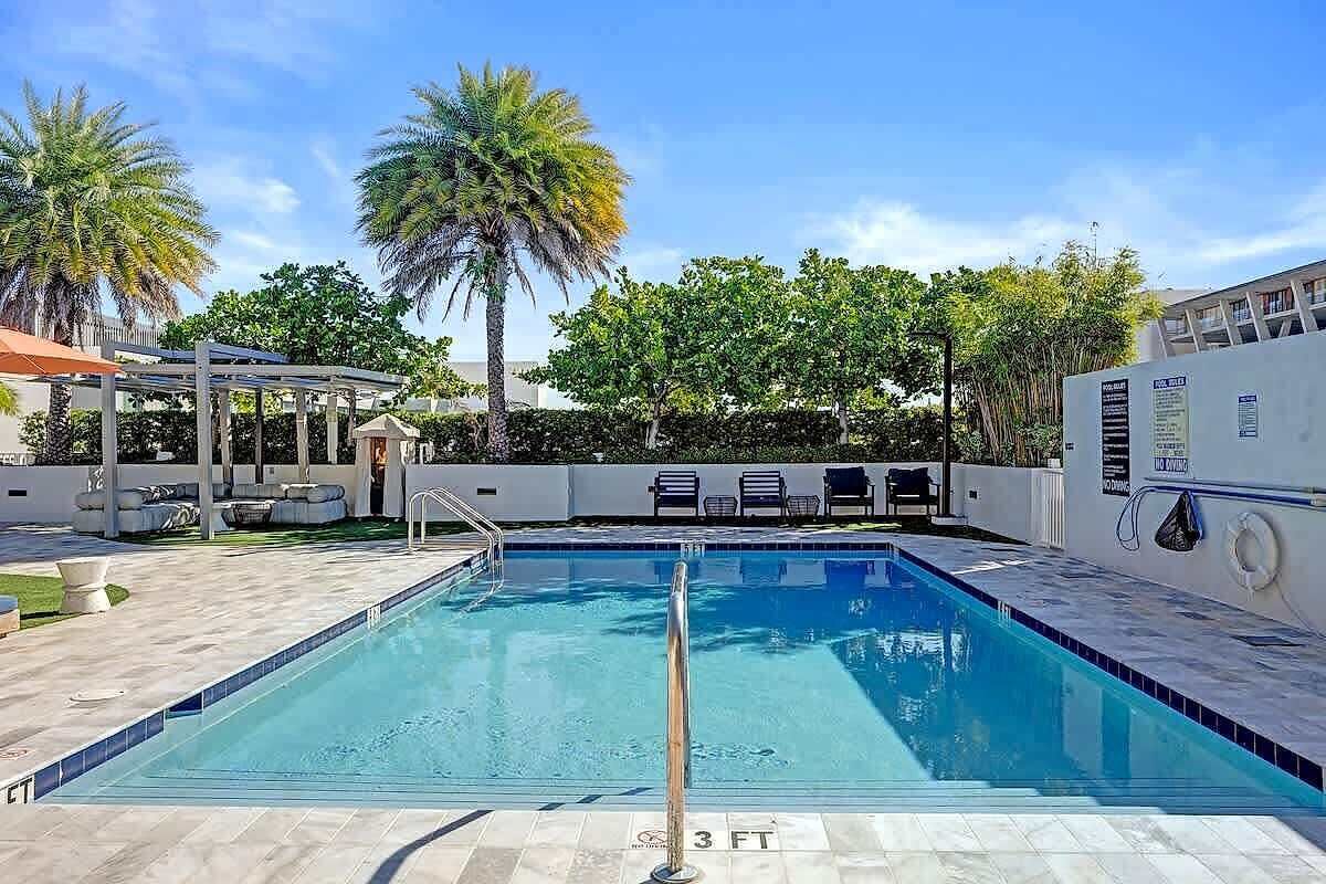 Ein rechteckiger Pool mit Metall-Sicherheitsgeländer, umgeben von einer gefliesten Terrasse, Palmen und einem überdachten Sitzbereich.
