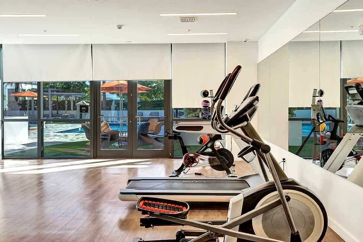 A gym with cardio equipment, large windows, and a view of outdoor seating.