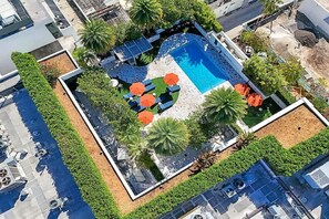 Vista aerea di una zona piscina su un tetto, con poltrone da relax e ombrelloni, circondata da vegetazione e palme.