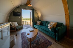 A cozy interior with a green sofa, a wooden table, and a bench. A television is mounted on the wall, and there's a view of the outdoors through a door.