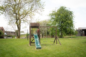 Une aire de jeux avec un toboggan et des balançoires, entourée d’herbe et d’arbres.