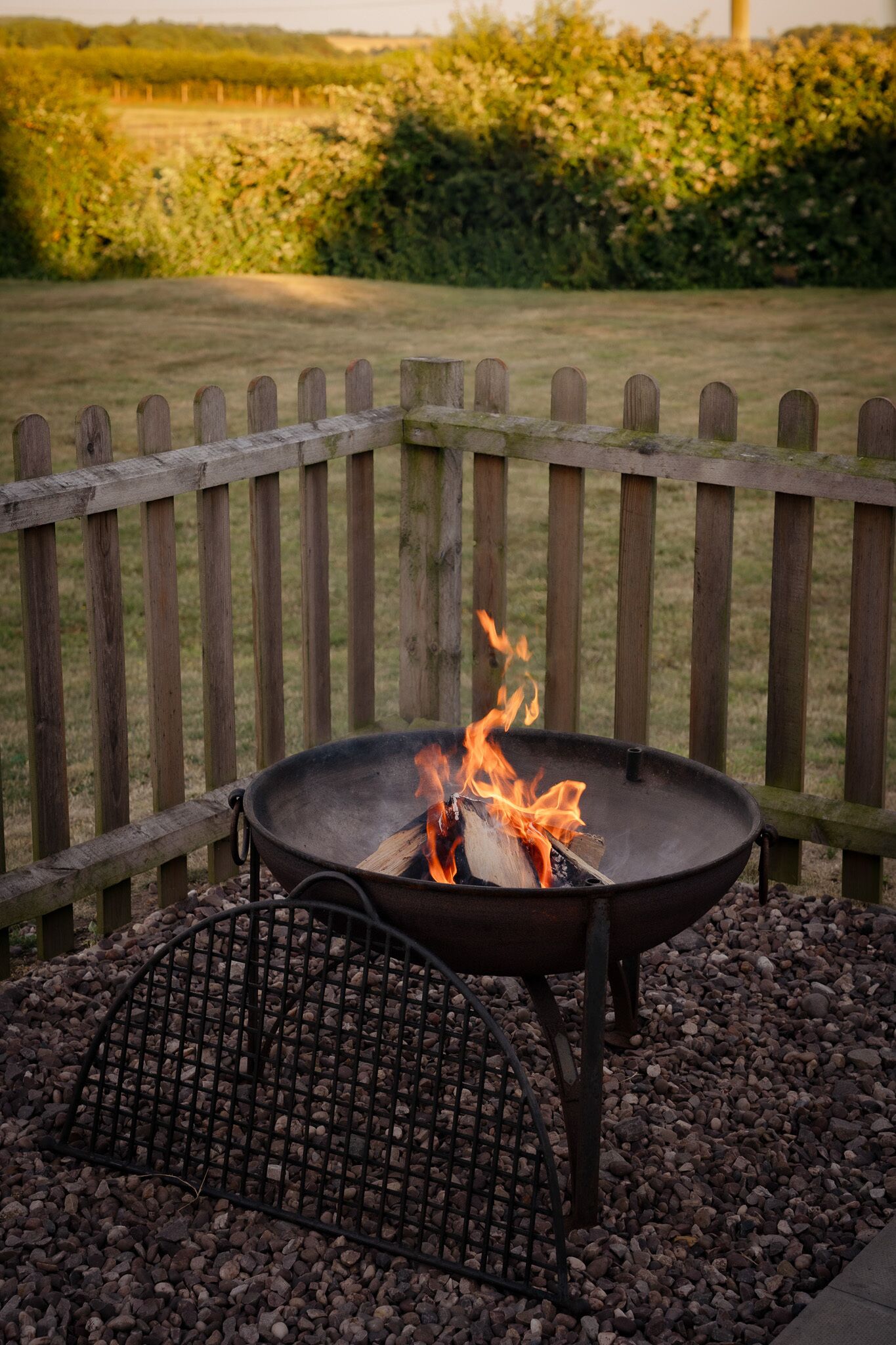 A metal fire pit with flames, surrounded by a wooden fence and gravel.