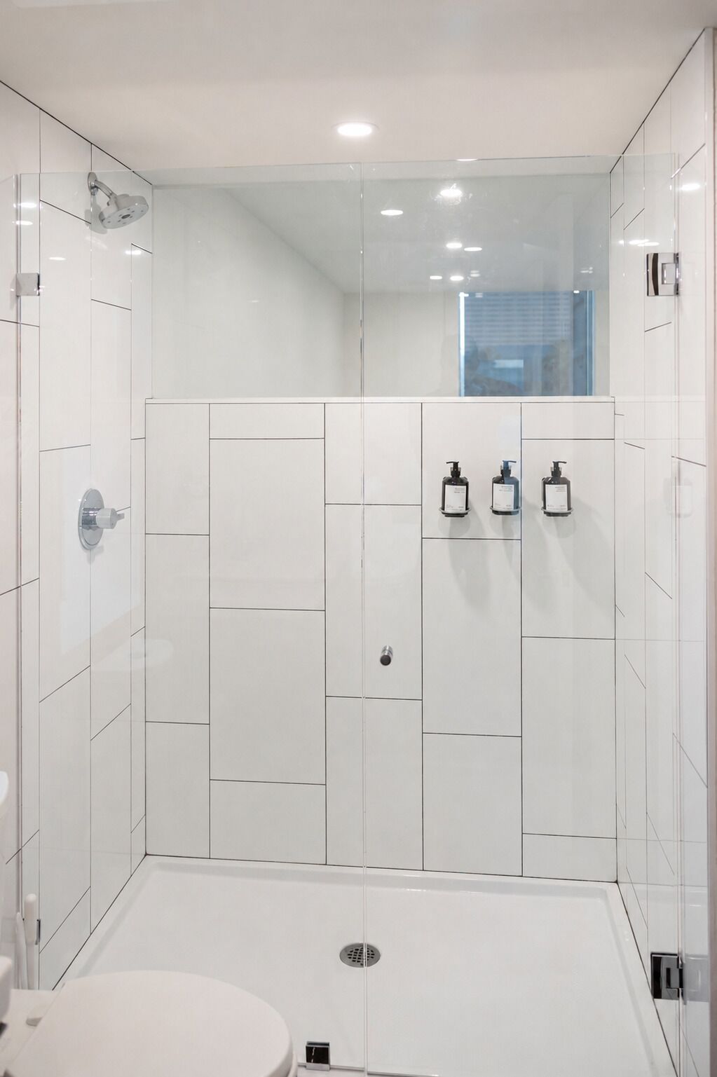 A modern bathroom with white tiles, a shower, and two hand soap dispensers on the wall.