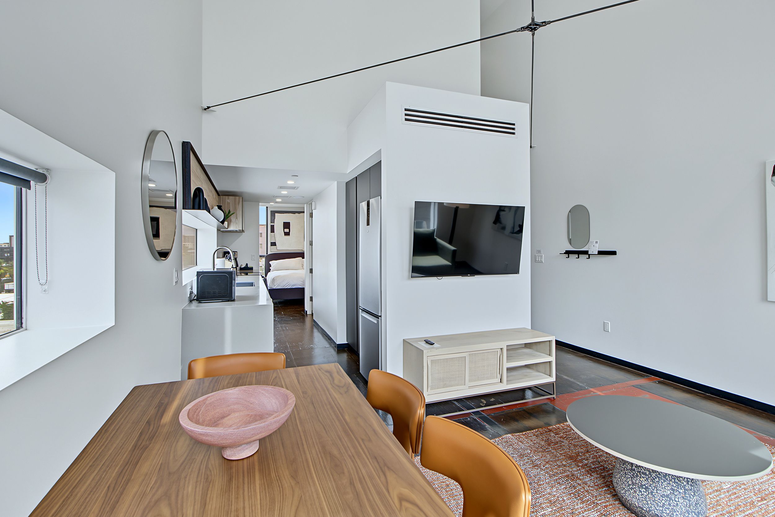 A modern hotel room with a wooden dining table, a television, and a kitchenette.