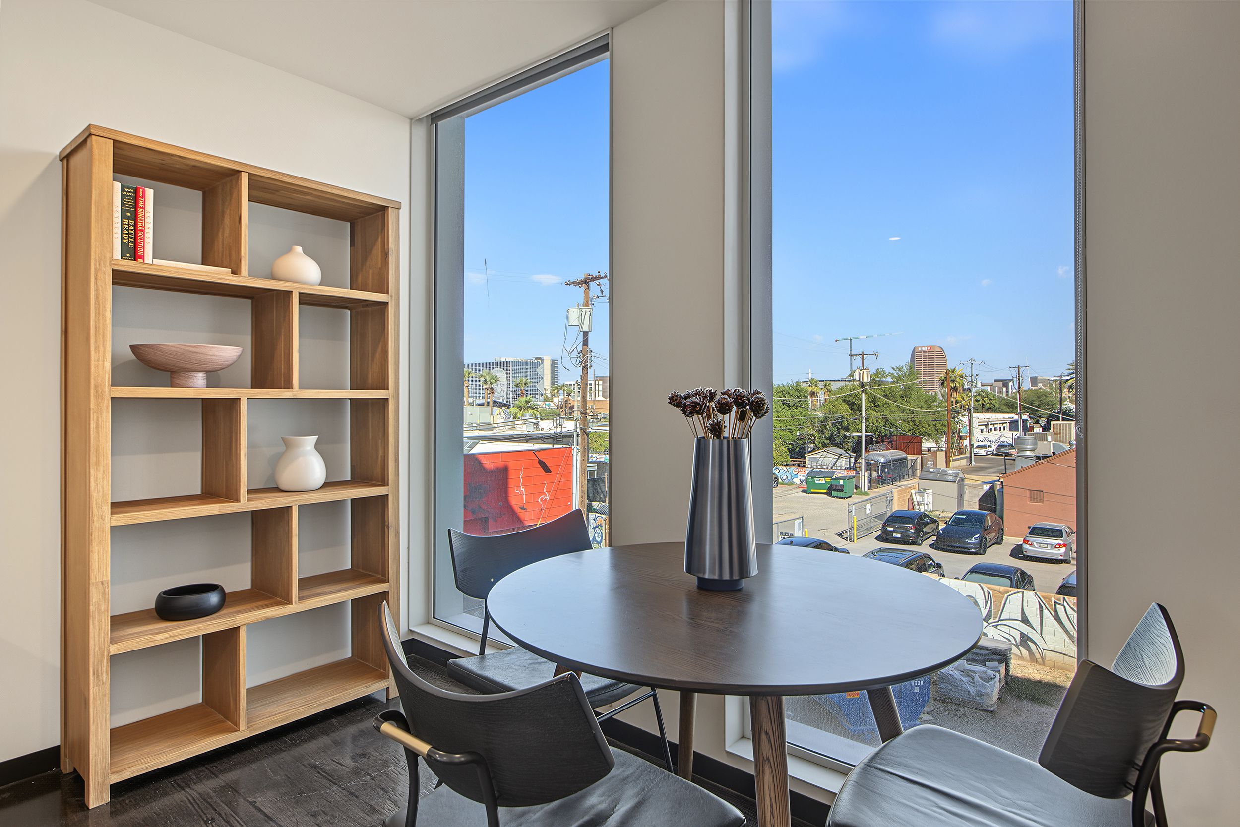 A room with a large window, a round table, two chairs, a bookshelf, and a vase with flowers.