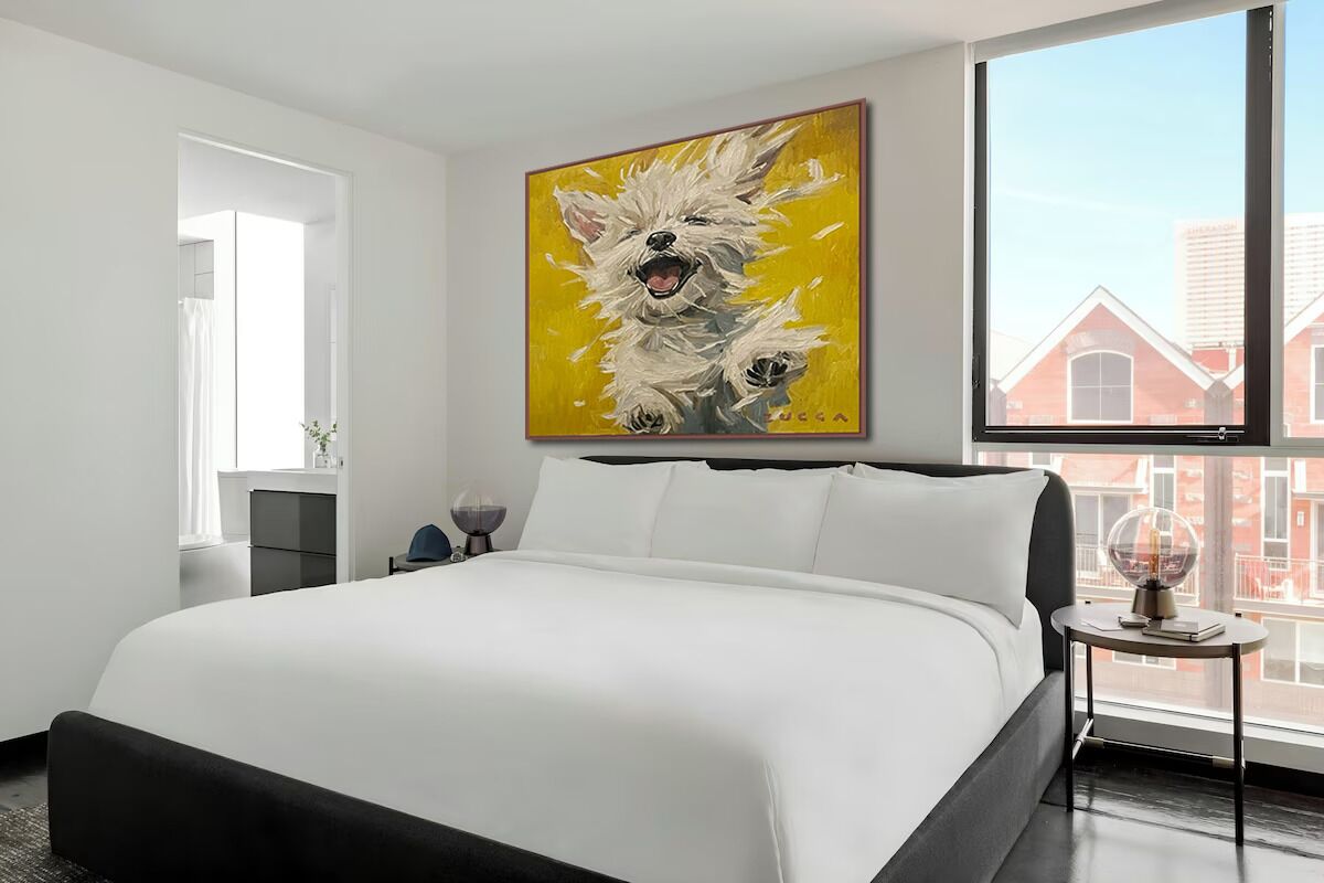 A modern hotel room with a large bed, a painting of dogs on the wall, a window with a view of buildings, and a small table with a lamp.