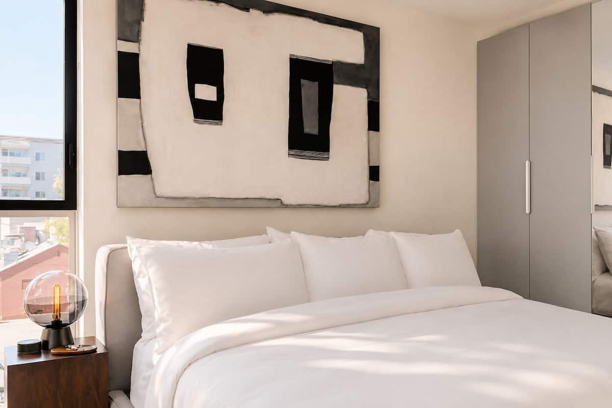 Un dormitorio con cama, mesita de noche, lámpara y una obra de arte en la pared.