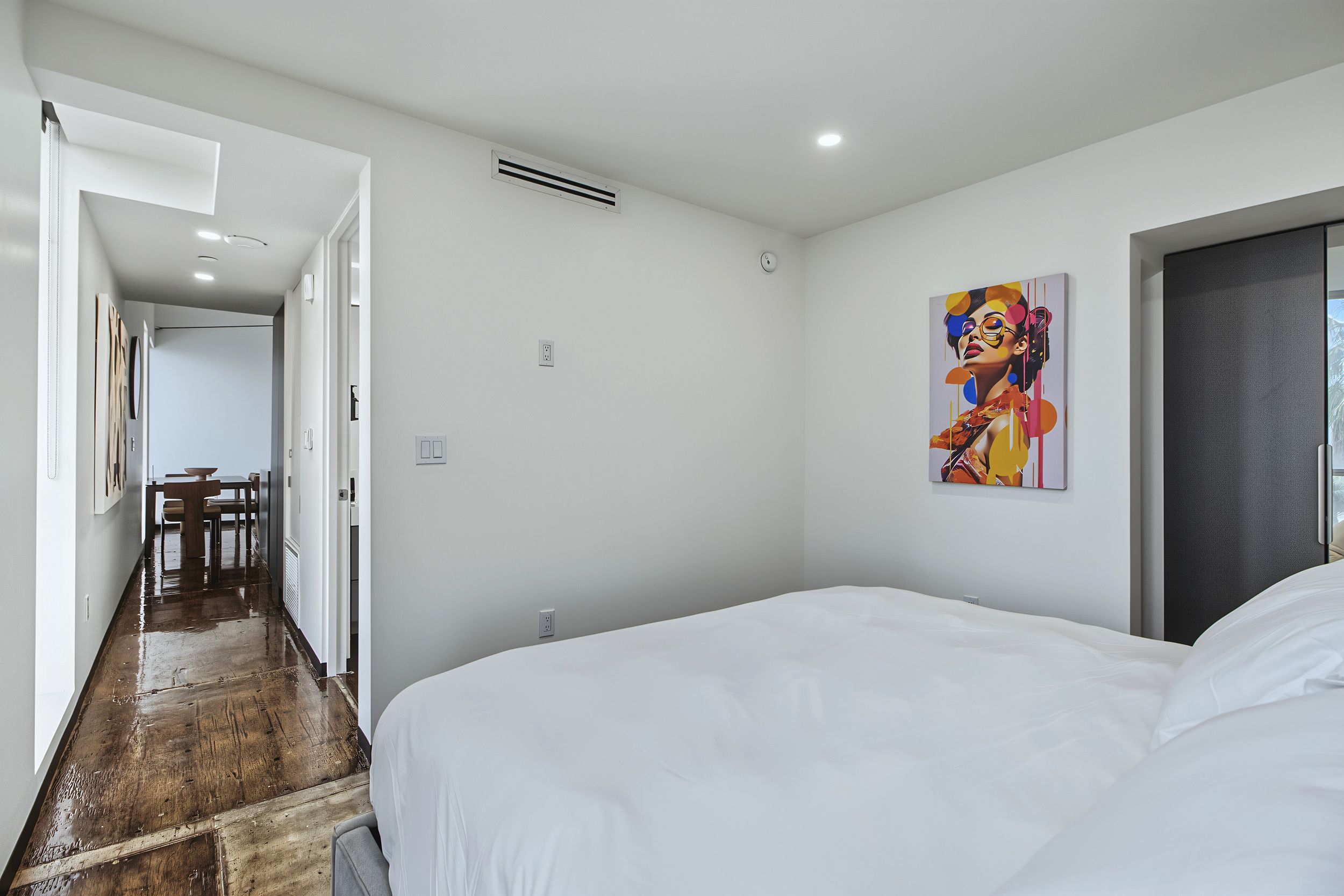 A hotel room with a large bed, a painting on the wall, and a hallway leading to a dining area.