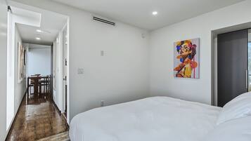 A hotel room with a large bed, a painting on the wall, and a hallway leading to a dining area.