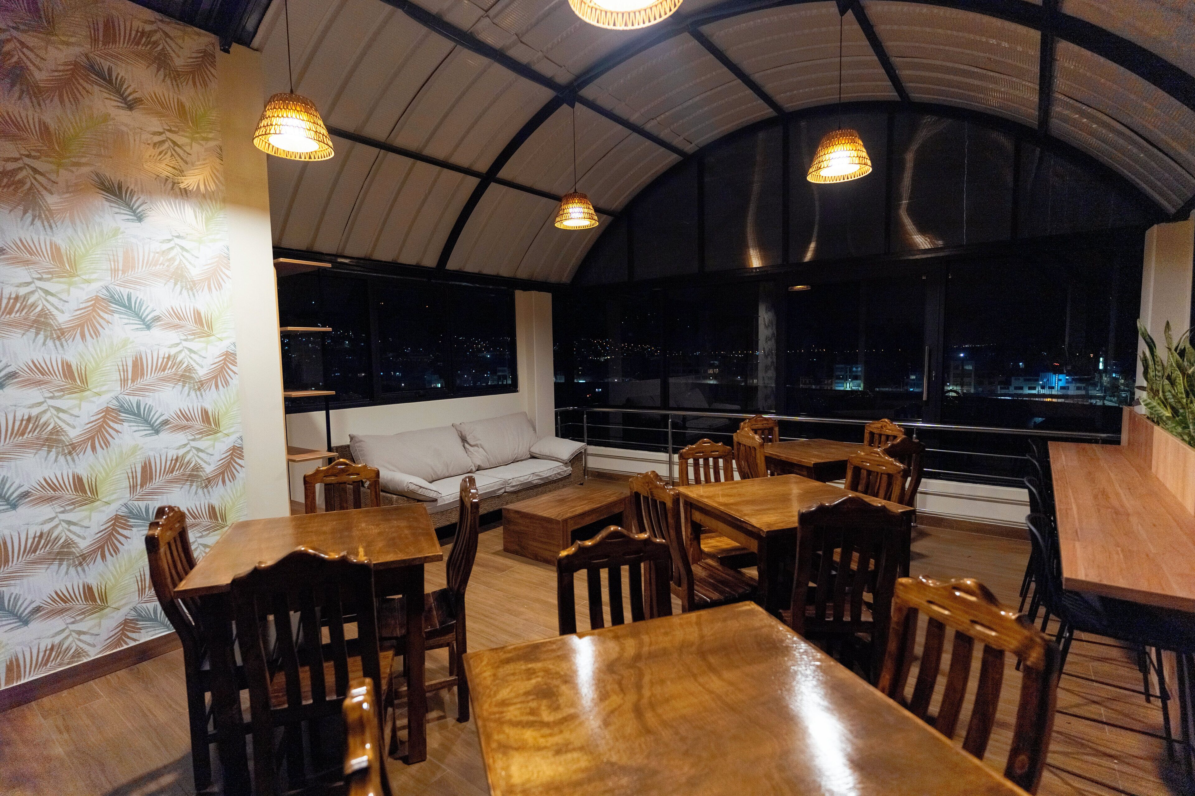 A restaurant with wooden tables and chairs, a sofa, and large windows with a city view at night.