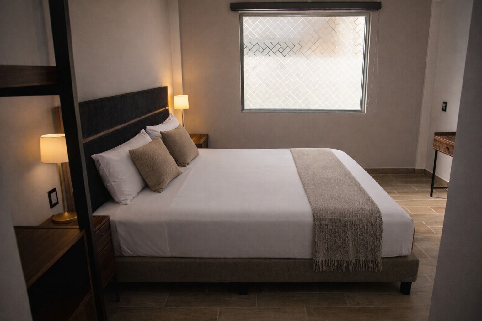 Una cama bien hecha con cabecera, dos lámparas de noche, una ventana con vistas y una mesita de noche de madera.