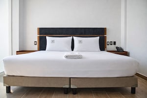 Una cama bien tendida con un cabecero oscuro, sábanas blancas y una toalla doblada. Hay dos mesitas de noche con lámparas y un teléfono.