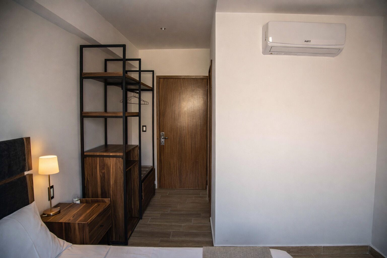 Une chambre d’hôtel avec un lit en bois, une table de chevet avec une lampe, une étagère intégrée et un climatiseur fixé au mur.