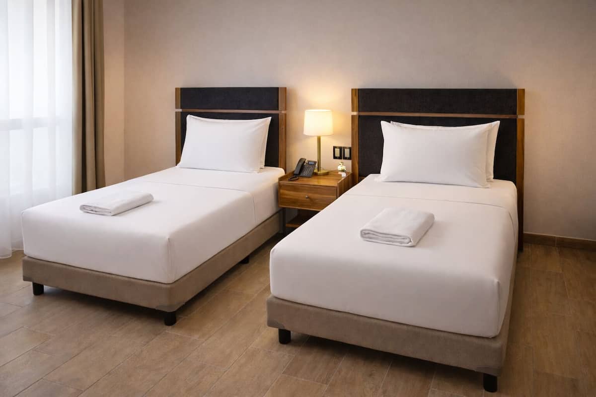 Deux lits simples avec du linge de lit et des serviettes blanches, une table de chevet avec une lampe, et un plancher en bois.
