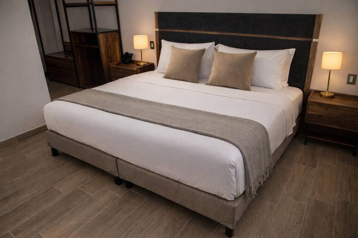 Un lit bien fait, recouvert d’un plaid gris, avec deux tables de chevet dotées de lampes et une tête de lit en bois.