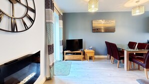 Ein modernes Wohnzimmer mit Kamin, Flachbildfernseher, Essbereich mit Glastisch und Ledersesseln sowie einer großen Wanduhr.