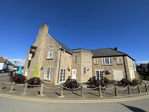A historic stone building with a prominent chimney, multiple windows, and a paved area with wooden chairs and tables.
