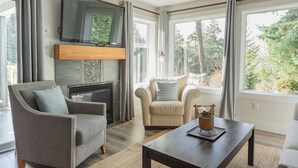 Una sala de estar con chimenea, dos sillones, una mesa de centro y ventanas grandes con cortinas.