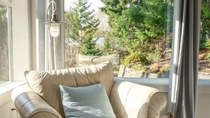 Un sillón de cuero color beige con un cojín azul claro, situado cerca de una ventana con vistas a árboles y una pared de piedra.