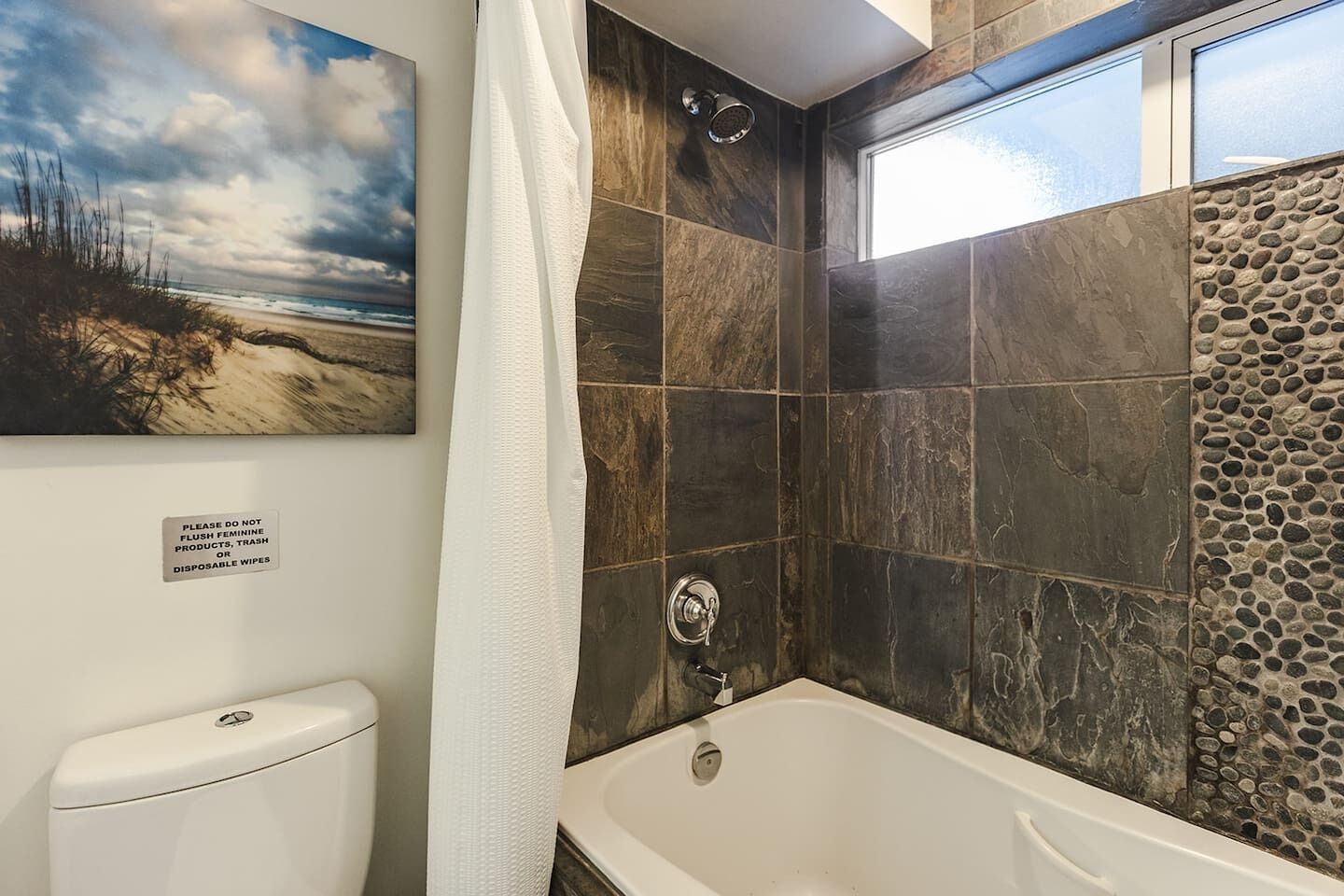 A bathroom with a bathtub, a shower, and a toilet. There is a sign on the wall and a painting above the toilet.
