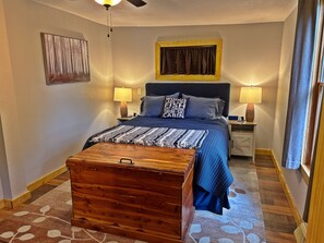 A bedroom with a bed, two bedside tables, a wooden chest, a ceiling fan, a painting on the wall, and a window with curtains.