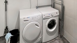 Deux machines à laver à chargement frontal se trouvent dans une buanderie dont les murs sont recouverts de carreaux et qui est équipée d’une étagère en métal.