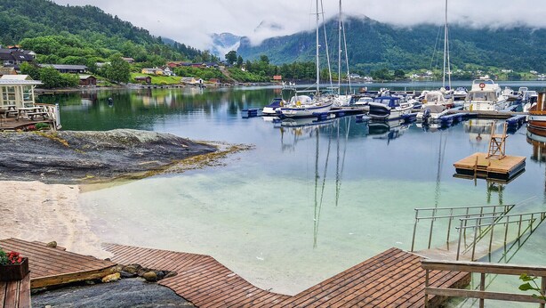 Uma marina com barcos atracados, um píer de madeira e montanhas ao fundo.
