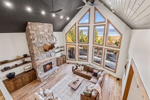 A spacious living room with a stone fireplace, wooden beams, and large windows offering a view of the outdoors.