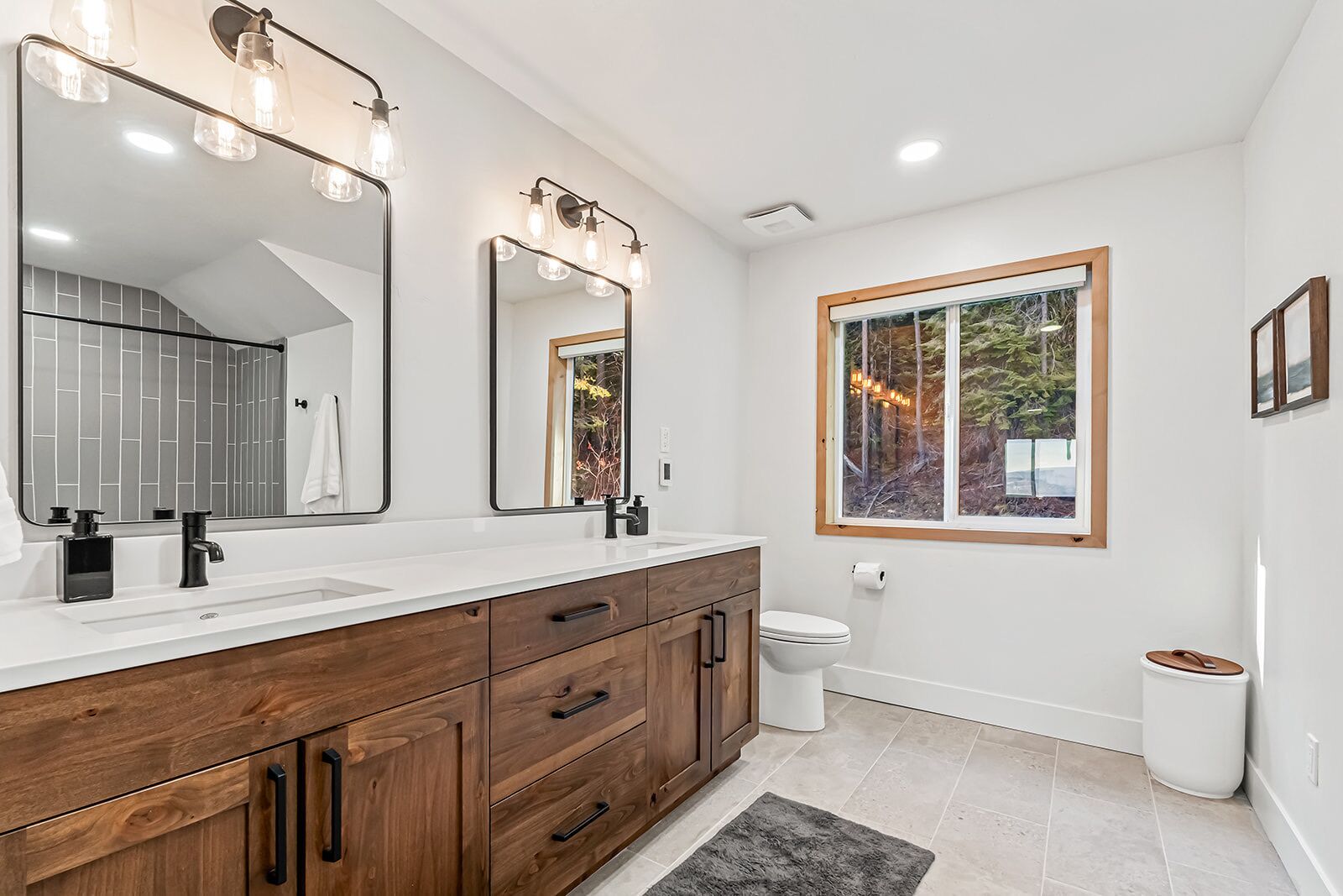 A modern bathroom with a double vanity, large mirrors, and a window with a view of trees.
