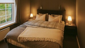 Cama bem arrumada com colcha xadrez e duas almofadas, com mesas de cabeceira e abajures de cada lado.