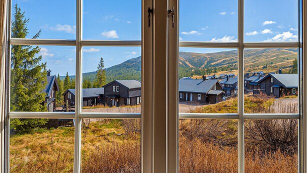 Vista pela janela de um edifício com vários quartos, uma porta e uma paisagem com casas e montanhas.