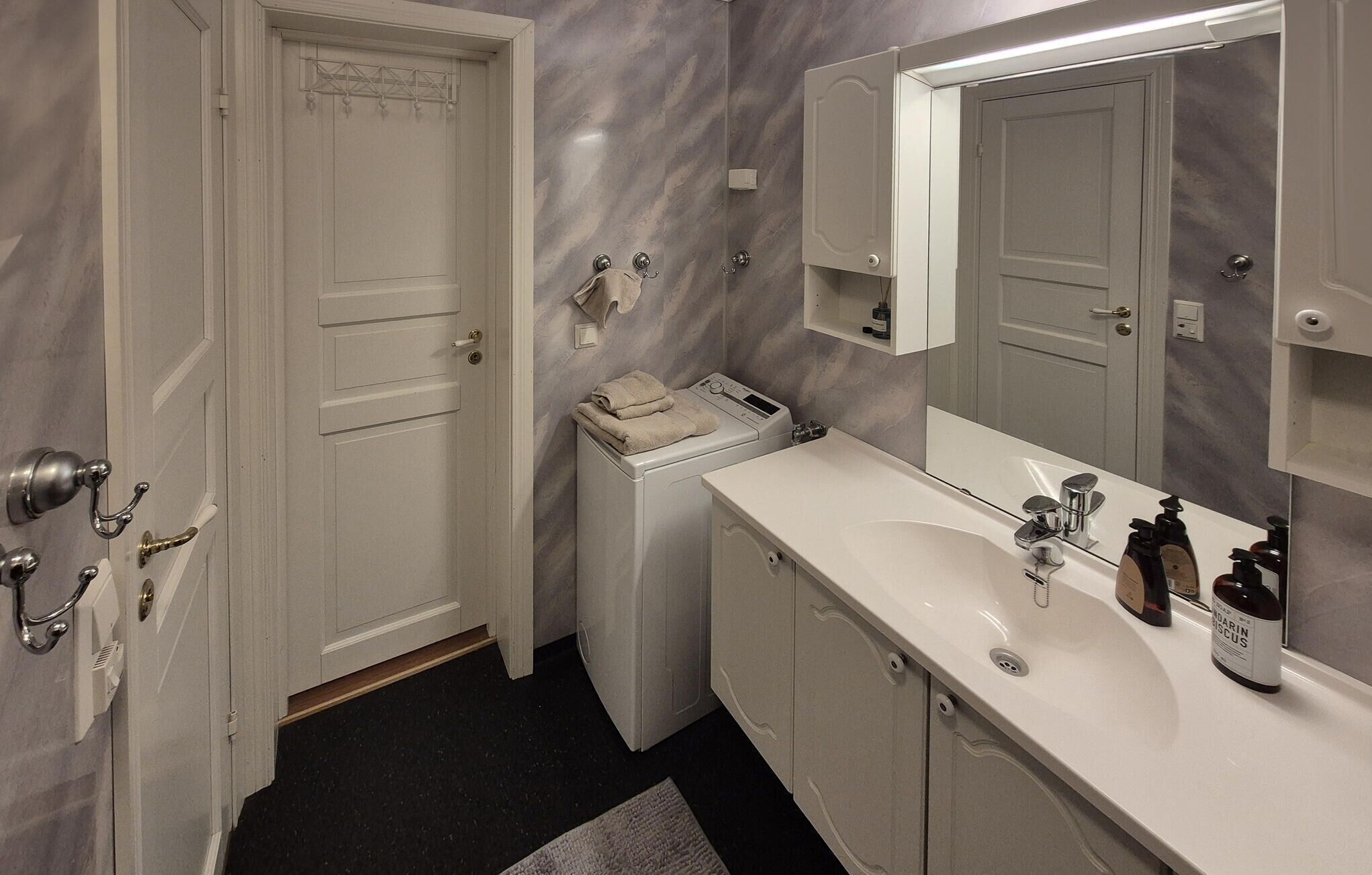 Une salle de bain avec un lavabo blanc, un miroir et un porte-serviettes. On y trouve également un lave-linge et une porte sur laquelle est accrochée une serviette.