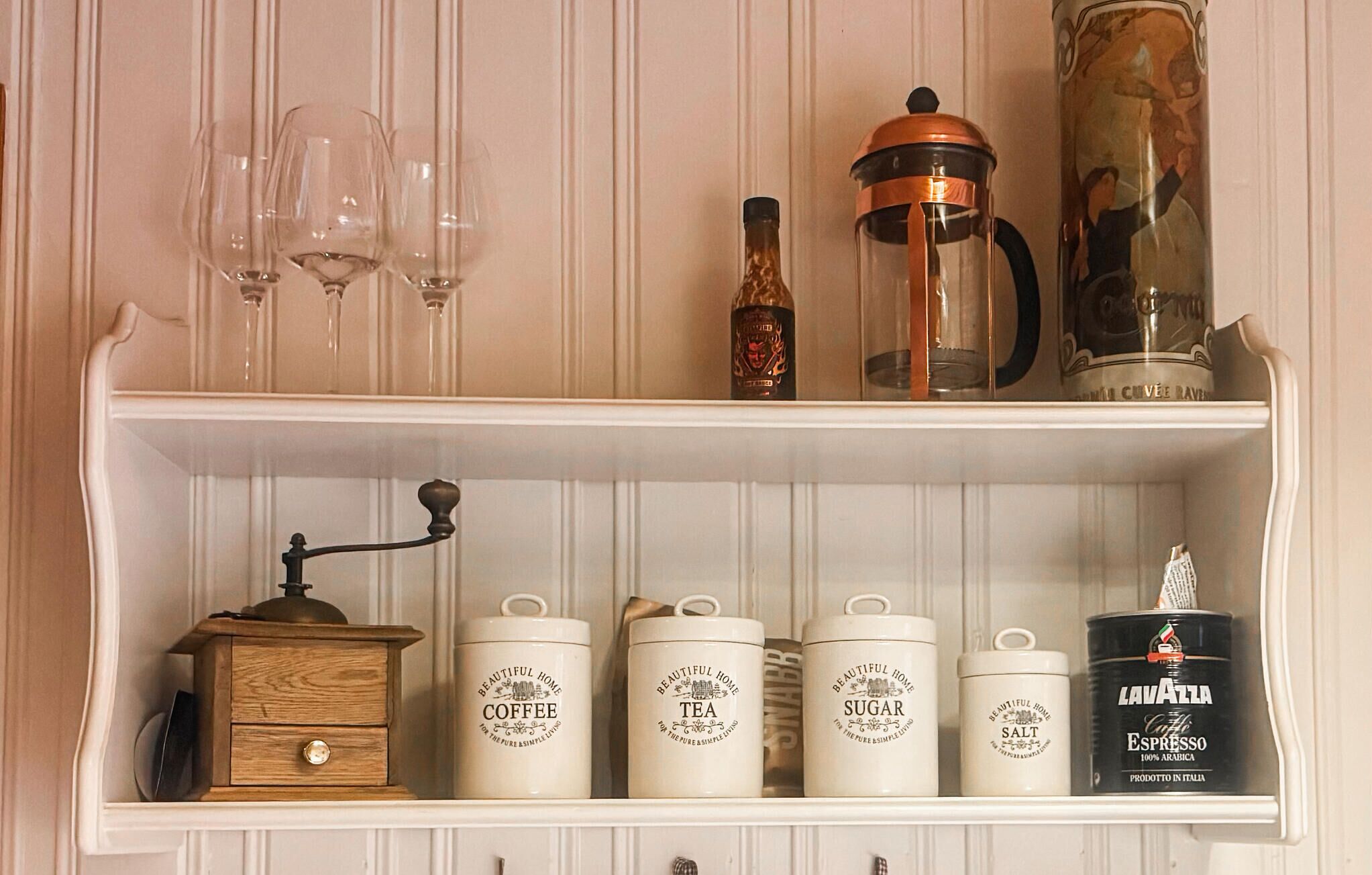 A shelf with various kitchen items including coffee, tea, sugar, salt containers, a coffee grinder, a French press, and a bottle of espresso.