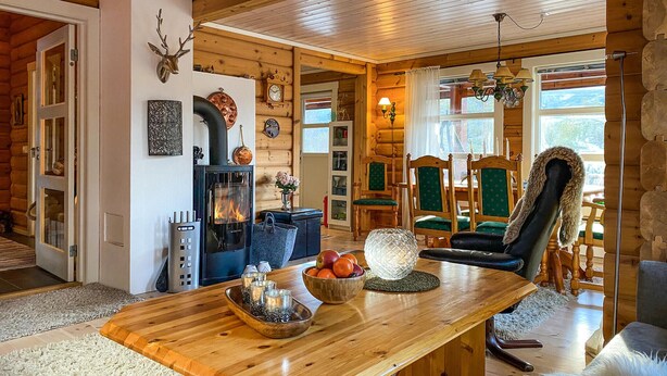 A cozy log cabin interior with a central wooden table, a fireplace, and a dining area with green upholstered chairs.