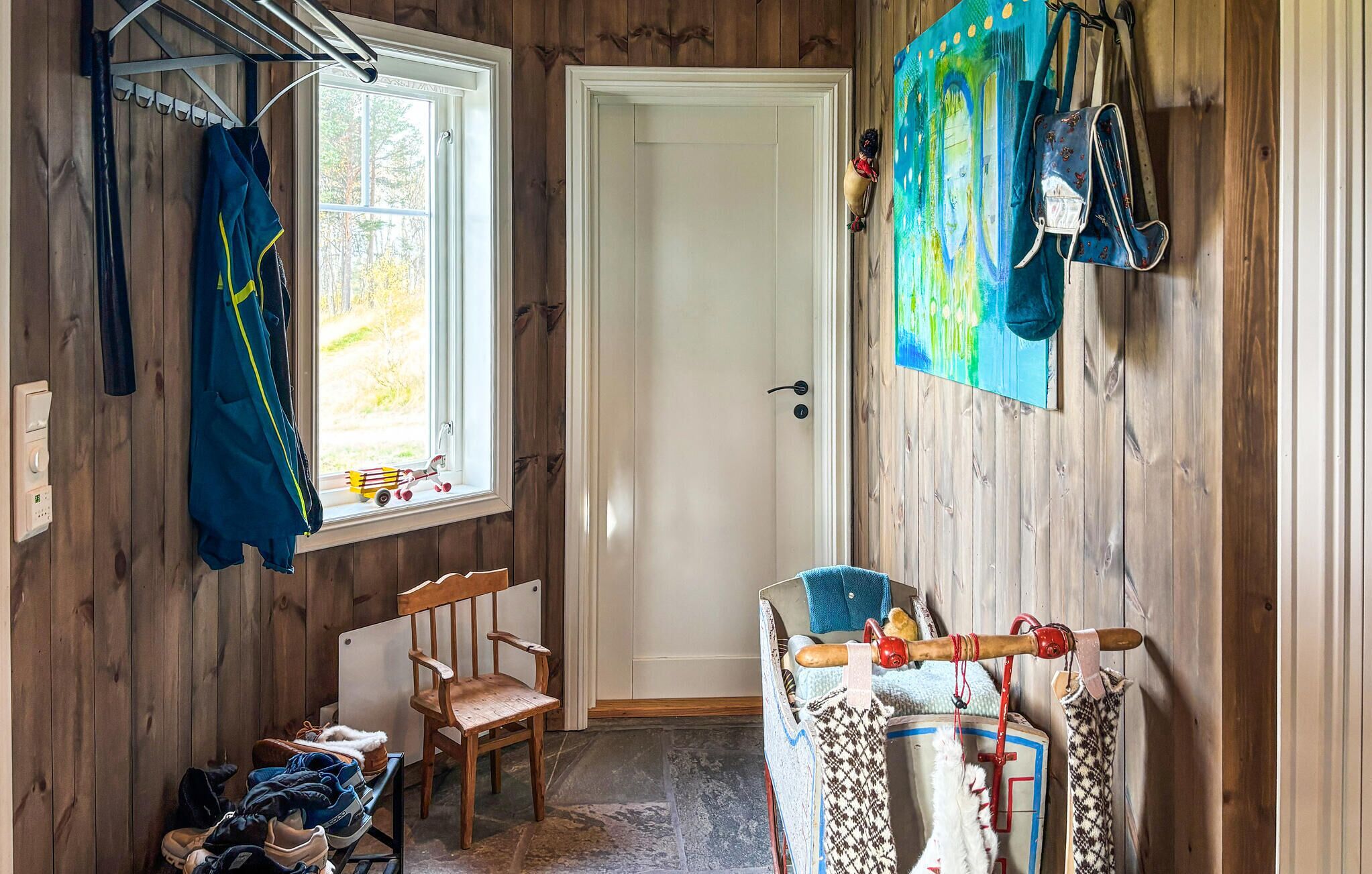 A room with wooden walls, a door, a window, a chair, a small table with shoes, and a hanging bag.