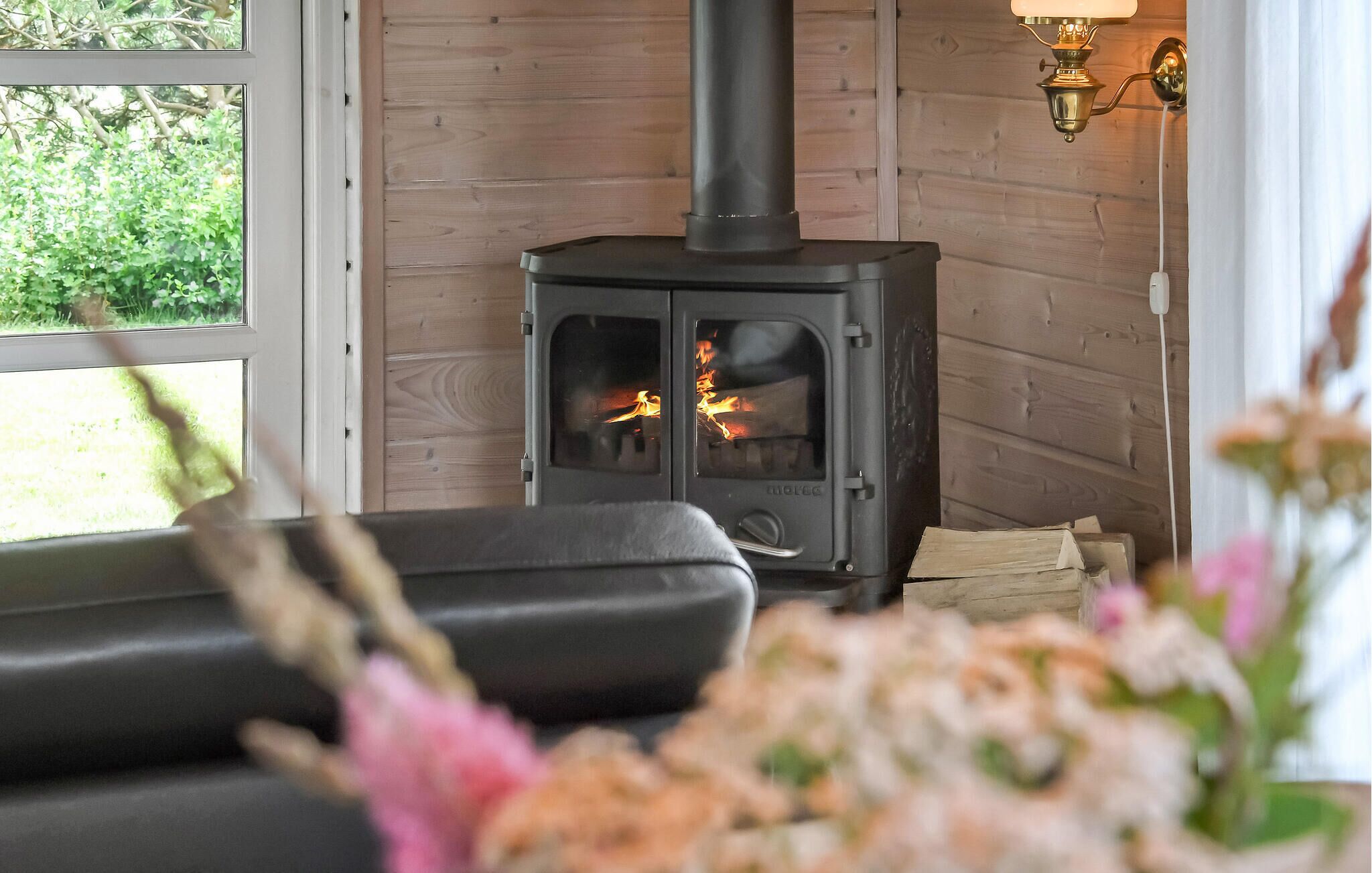 A cozy room with a wood-burning stove, a window with a view of greenery, and a wall-mounted light fixture.