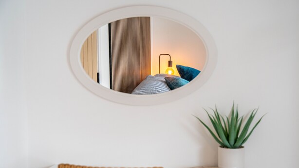 Espelho redondo refletindo um quarto com abajur, almofadas e planta.