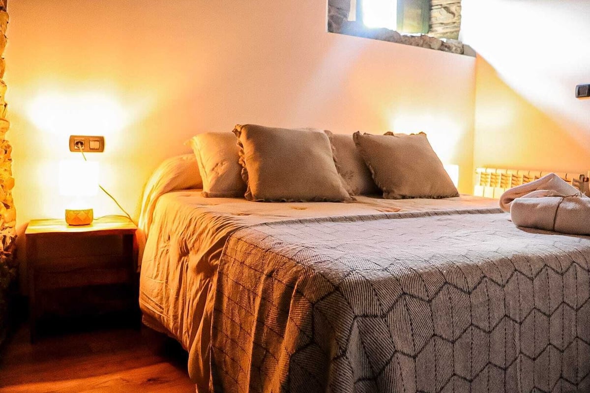 A bedroom with a bed, pillows, a nightstand, a lamp, and a window.