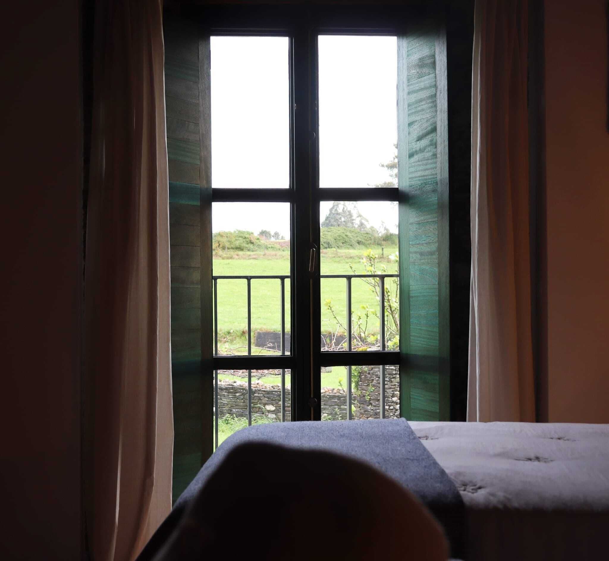 A room with a bed, a window with a view of a green landscape, and curtains.