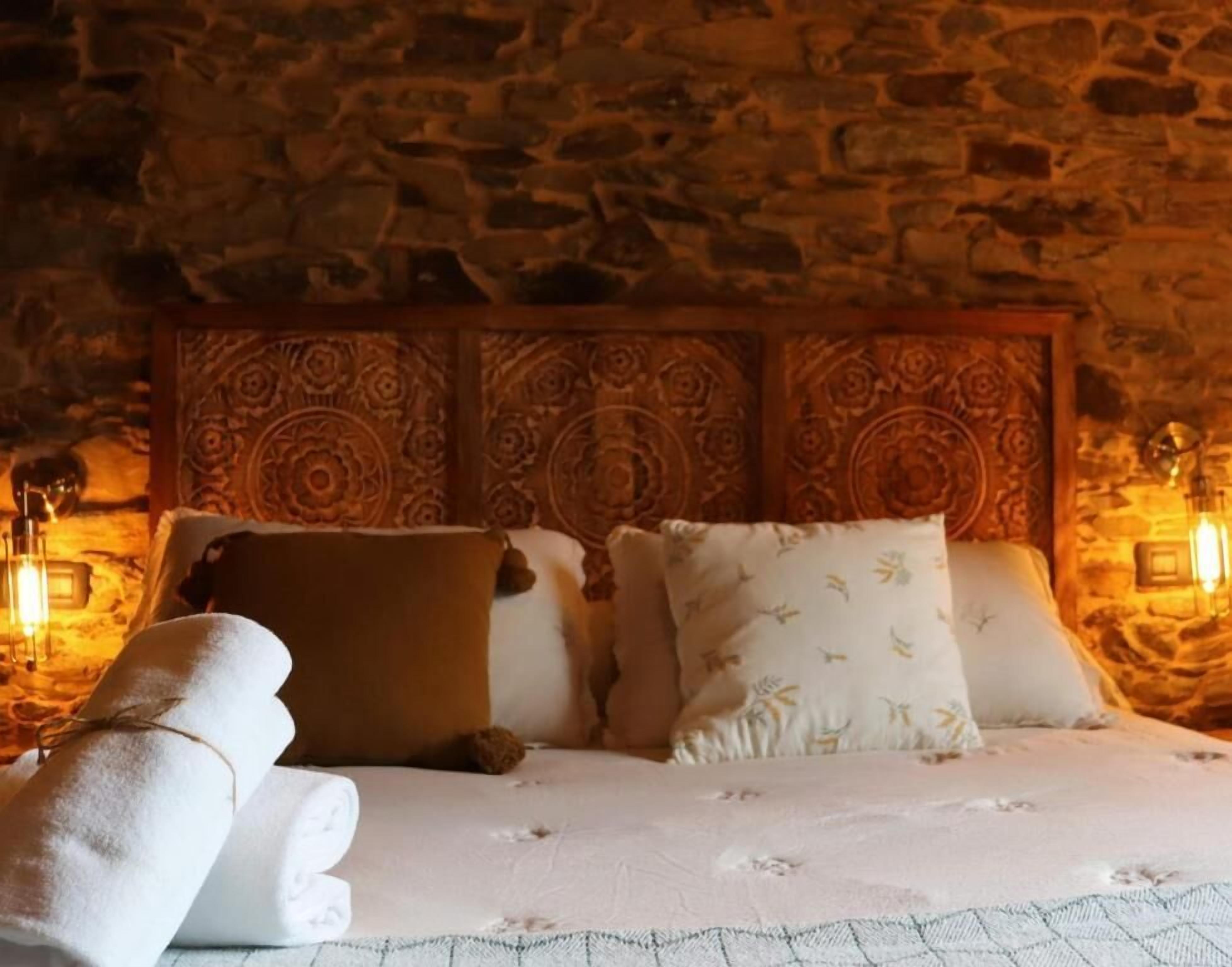 Una habitación acogedora con pared de piedra, cabecera de madera con intrincados grabados, y una cama con sábanas blancas y almohadas estampadas.