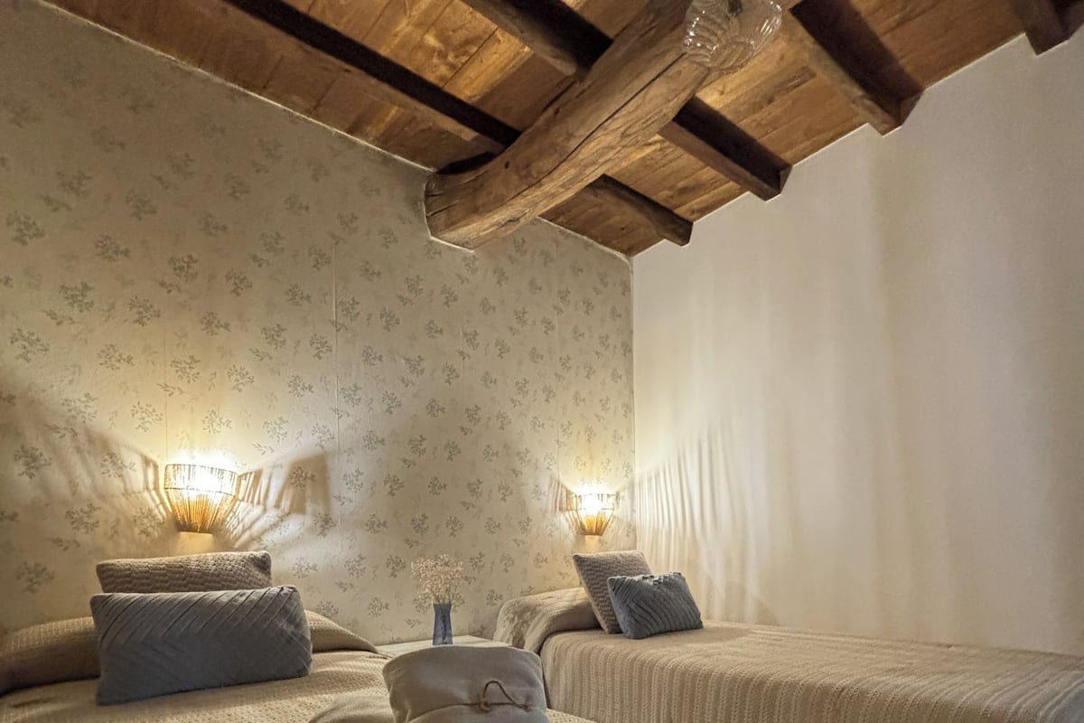 A cozy bedroom with two beds, wooden beams, floral wallpaper, and two wall-mounted lamps.