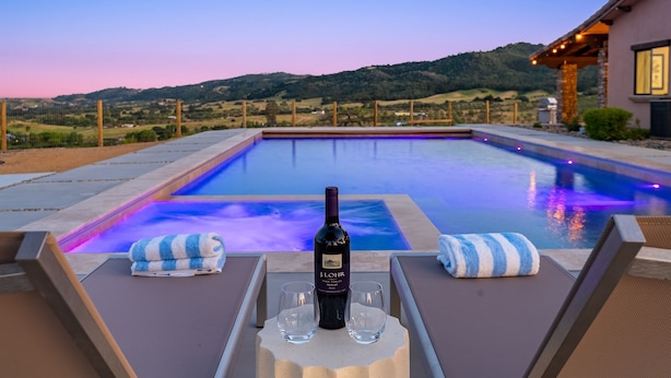 Área da piscina com espreguiçadeiras, uma garrafa de vinho e copos em cima de uma mesa, com vista para a piscina e colinas.
