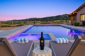 A poolside area with lounge chairs, a bottle of wine, and glasses on a table, overlooking a pool with a view of hills. - Villa Encanto: 7BR Estate, Heated Pool & Spa, Views (Paso Robles)