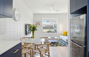 A modern kitchen with white tiles, a round dining table, wooden chairs, a refrigerator, and a window with a view of the outside. - Belle Escapes - Coral Drift Retreat (West Beach)