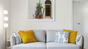 Ein modernes Wohnzimmer mit einem grauen Sofa, gelben und gemusterten Kissen, einer Stehlampe und einem gerahmten Bild von Pflanzen an der Wand.